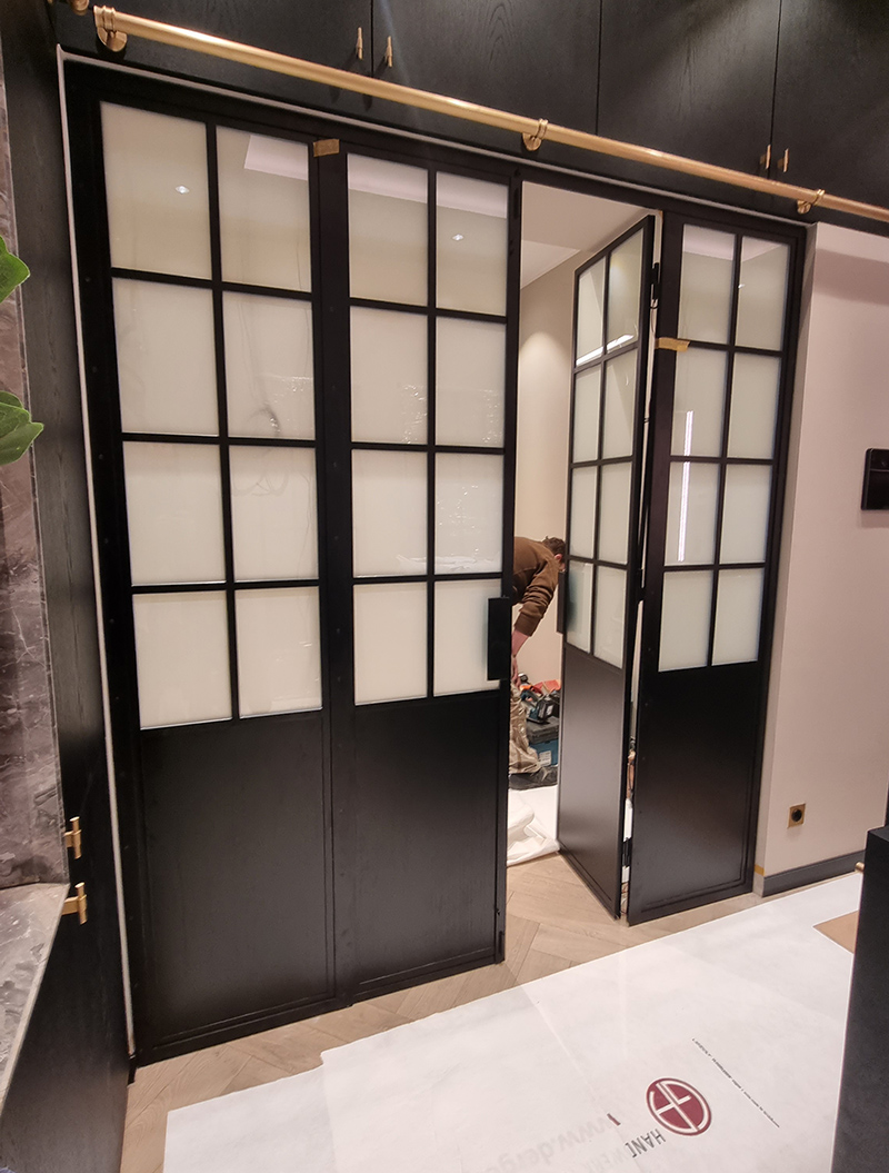 Moderne Glastür mit schwarzem Rahmen, Person arbeitet im Hintergrund, neutrale Farben, klare Linien.