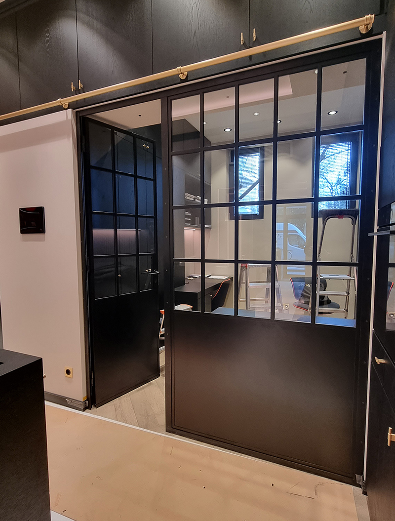Schwarze Schiebetür mit Glas, dahinter ein Raum mit Holz- und Metallmöbeln, weiße Wände, Tageslicht.