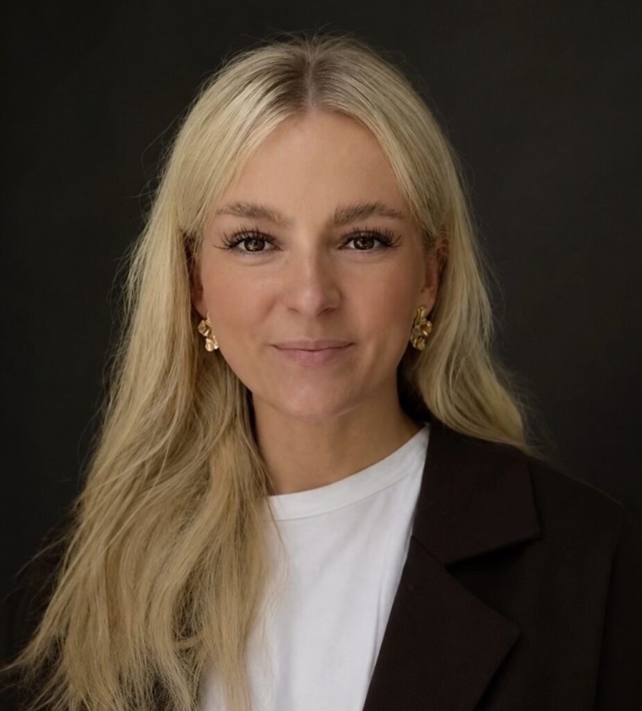 Blonde Frau mit sanftem Lächeln, schwarzem Blazer, goldenen Ohrringen