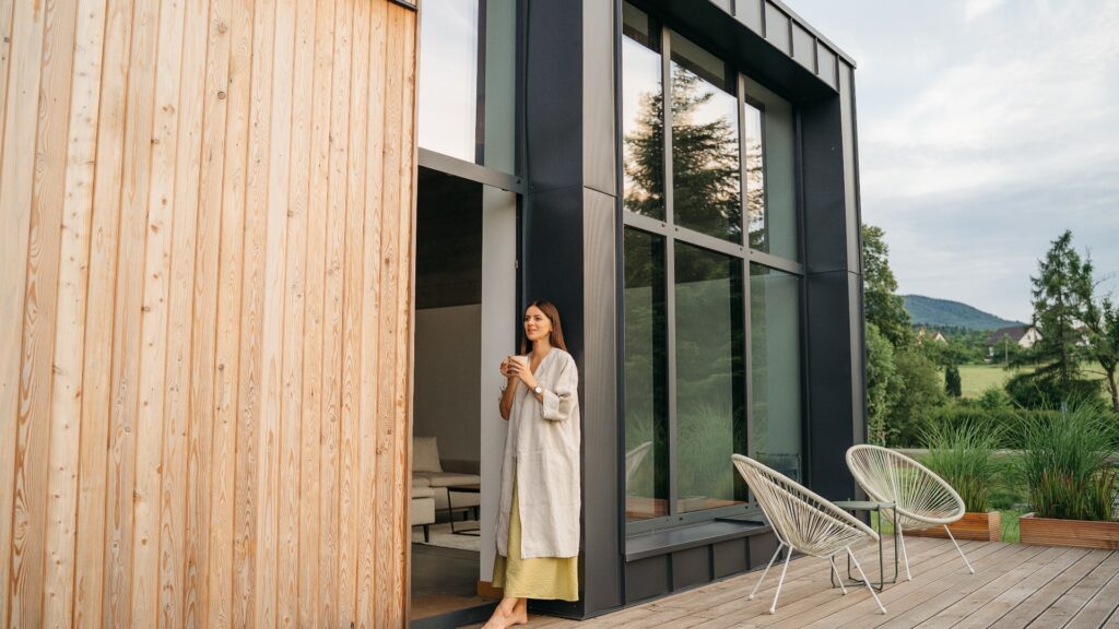 Eine junge Frau auf der Terrasse eines modernen Holzhauses mit Panoramafenstern.